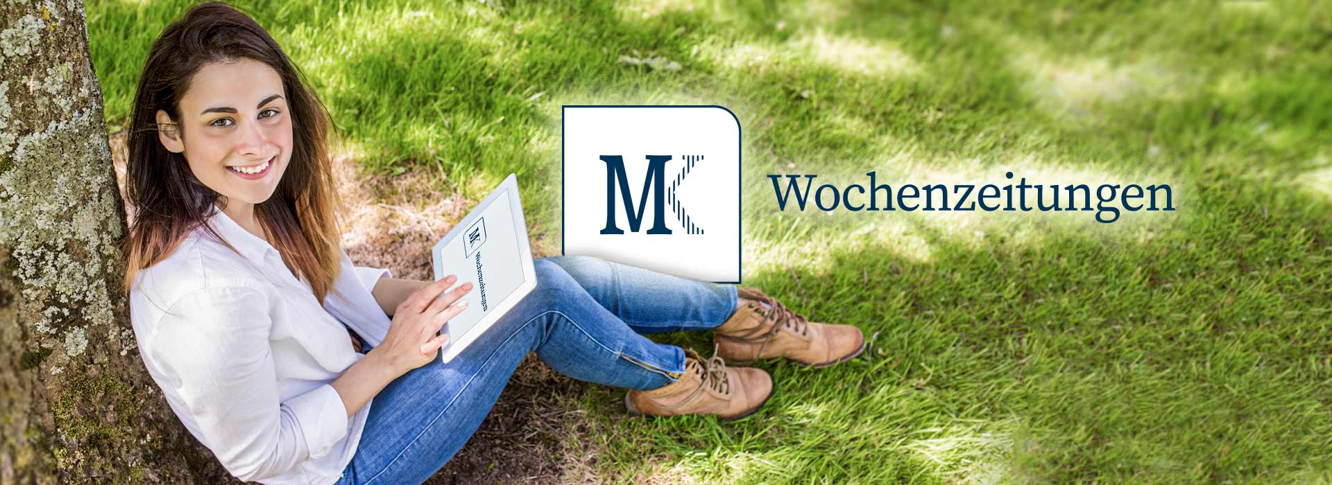 Frau sitzt auf Rasen und lehnt sich an einen Baum. Dabei hält sie ein Tablet mit dem MK Logo in der Hand